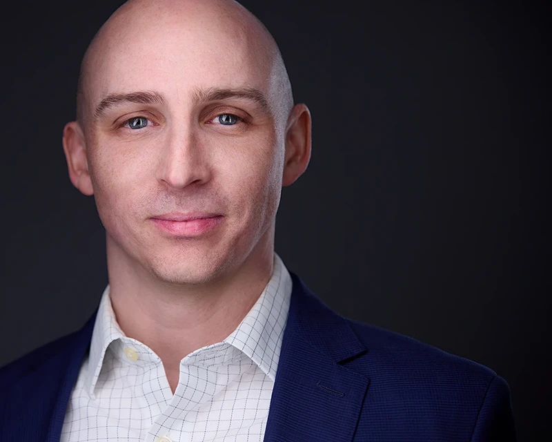 grinning headshot of white male in white shirt and blue suit on dark background