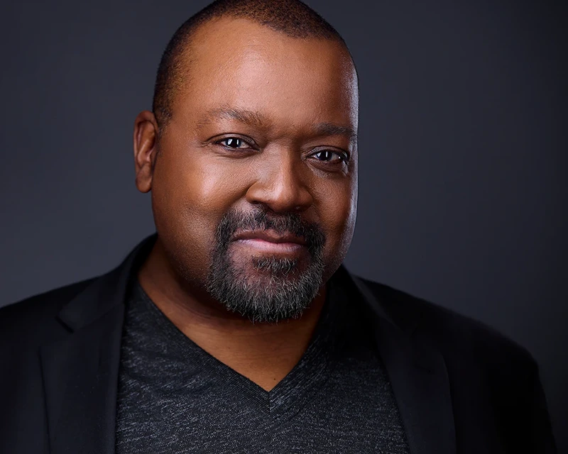 Confident black businessman with a wide, friendly grin, wearing a sleek black suit that blends seamlessly with the dark background, highlighting his warm expression.