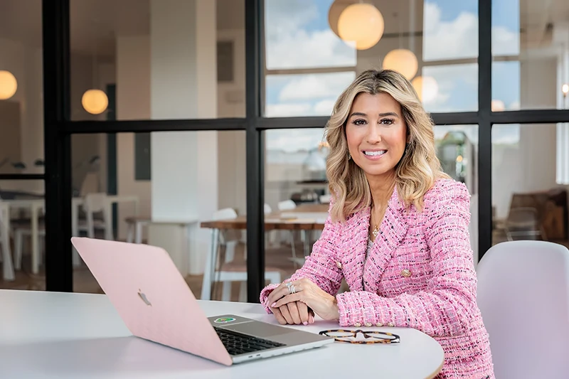 woman in pink suit sitting at a table with open laptop in a coworking space