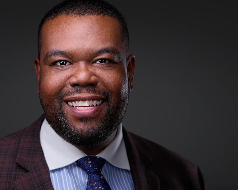 Grinning headshot of a black male dressed in a tailored brown suit and coordinating tie, showcasing approachability and charm.