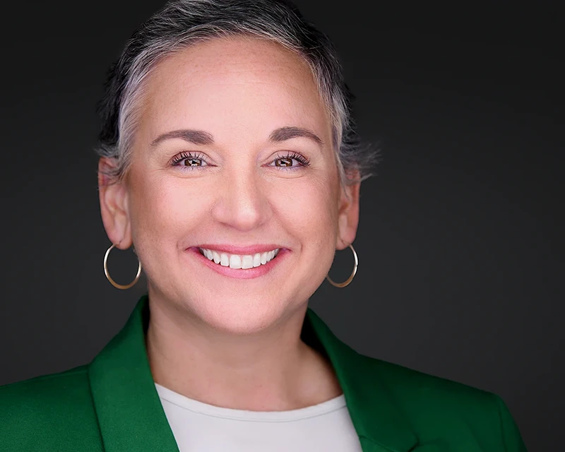 Striking headshot of a white woman with short gray hair, dressed in a bright green blazer, radiating professionalism and confidence in her appearance.