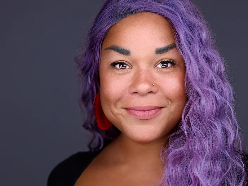 Stunning headshot of a confident black woman with vibrant purple hair, showcasing her unique style and personality against a soft background.