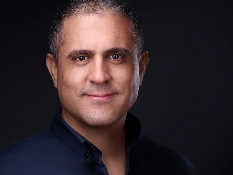 Professional headshot of a Latino man with short hair, dressed in a navy shirt, set against a sleek black background, conveying confidence and approachability.