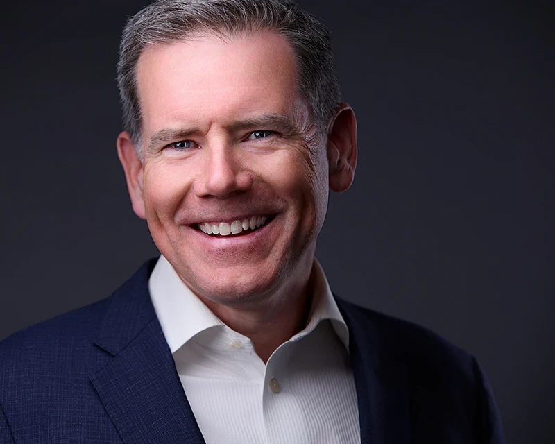 A professional headshot of a white man in a blue suit, smiling confidently against a soft gray backdrop, representing Pittsburgh's business community.