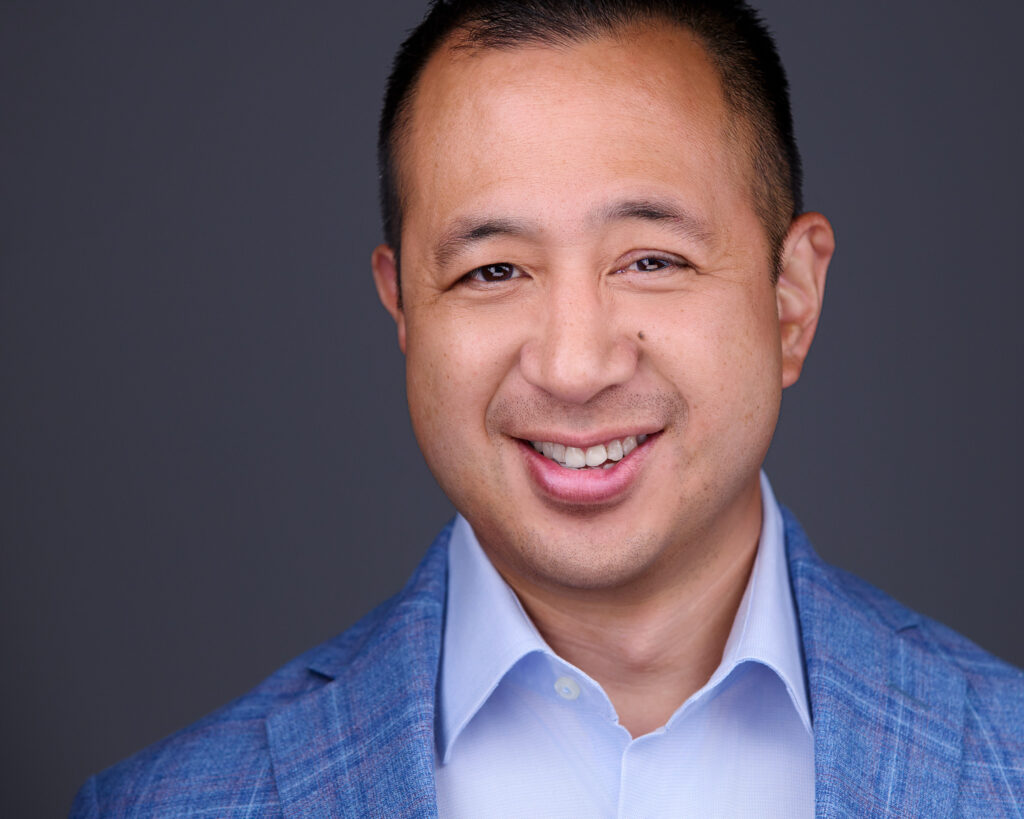 Smiling Asian man smiling with teeth for a LinkedIn headshots while wearing a blue shirt and blue plaid jacket