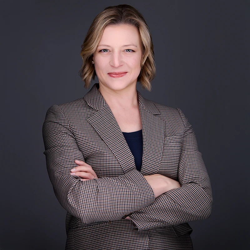 Self-assured female leader in sharp suit jacket stands with crossed arms, her infectious grin reflecting career satisfaction and expertise